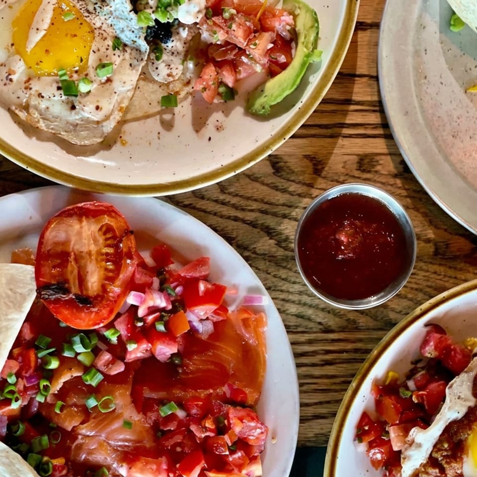 Overhead spread of breakfast plates with fried eggs, tortillas, avocado, tomatoes, salsa, and a small bowl of red sauce on a wooden table.