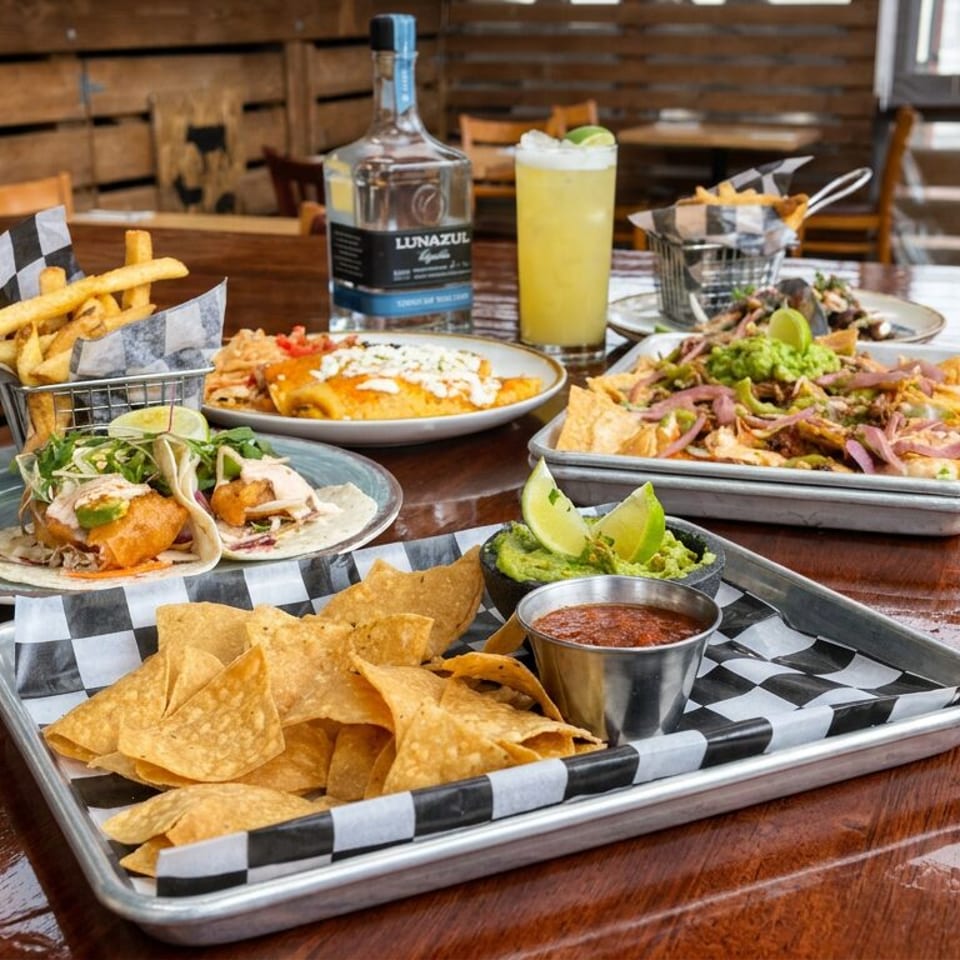 Mexican restaurant table featuring tacos, loaded fries, tortilla chips with guacamole and salsa, cocktails, and a tequila bottle.