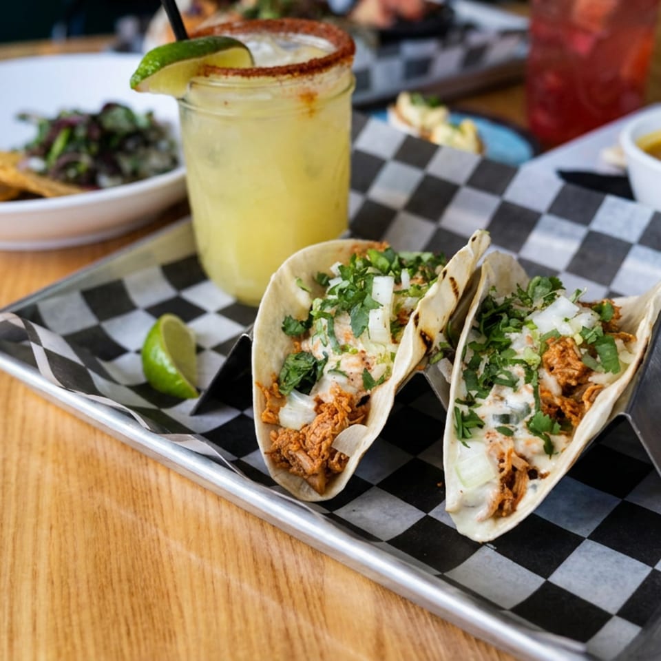Two cilantro-topped tacos with creamy sauce sit on a checkered tray alongside lime, chips, salsa, and a citrus drink.