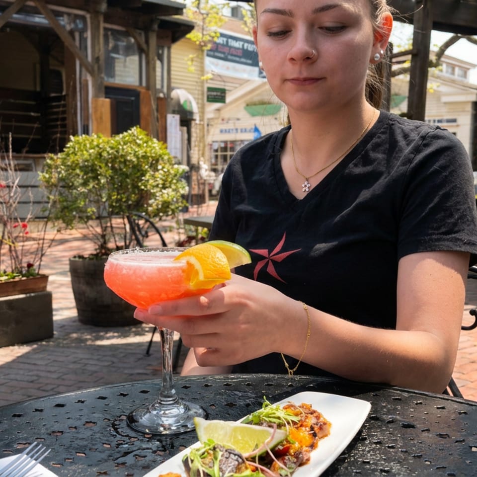 Server holds a bright pink citrus cocktail beside a plate of tacos on a sunny restaurant patio.
