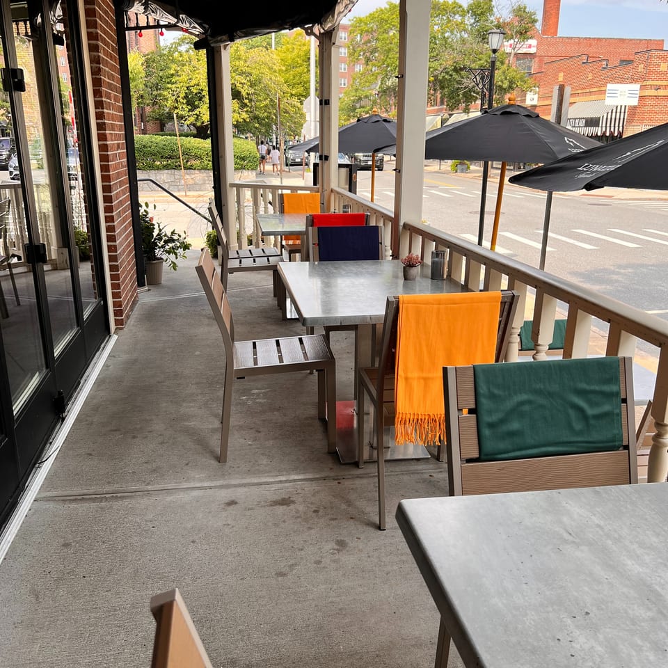 Covered restaurant patio with metal tables, colorful chairs, black umbrellas, and a sunny urban street view.