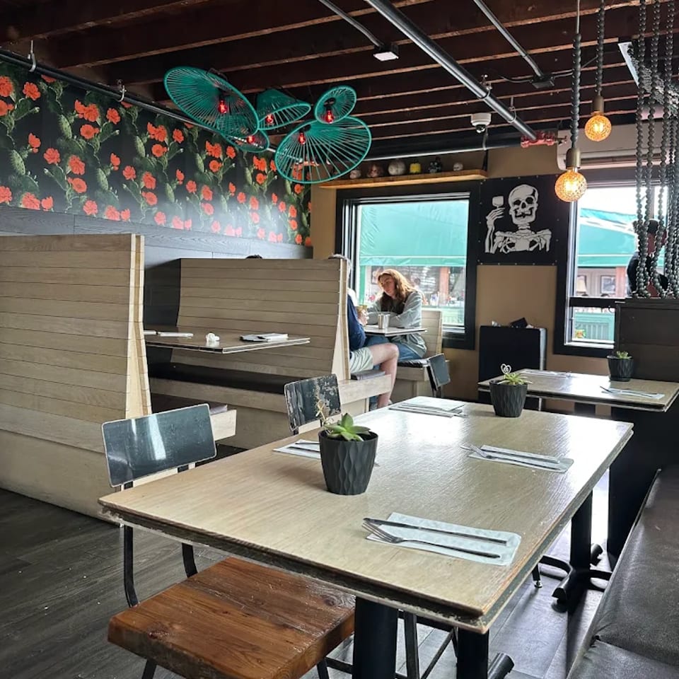 Eclectic restaurant interior with wooden tables, booth seating, floral wallpaper, hanging fans, plants, and skeleton artwork.