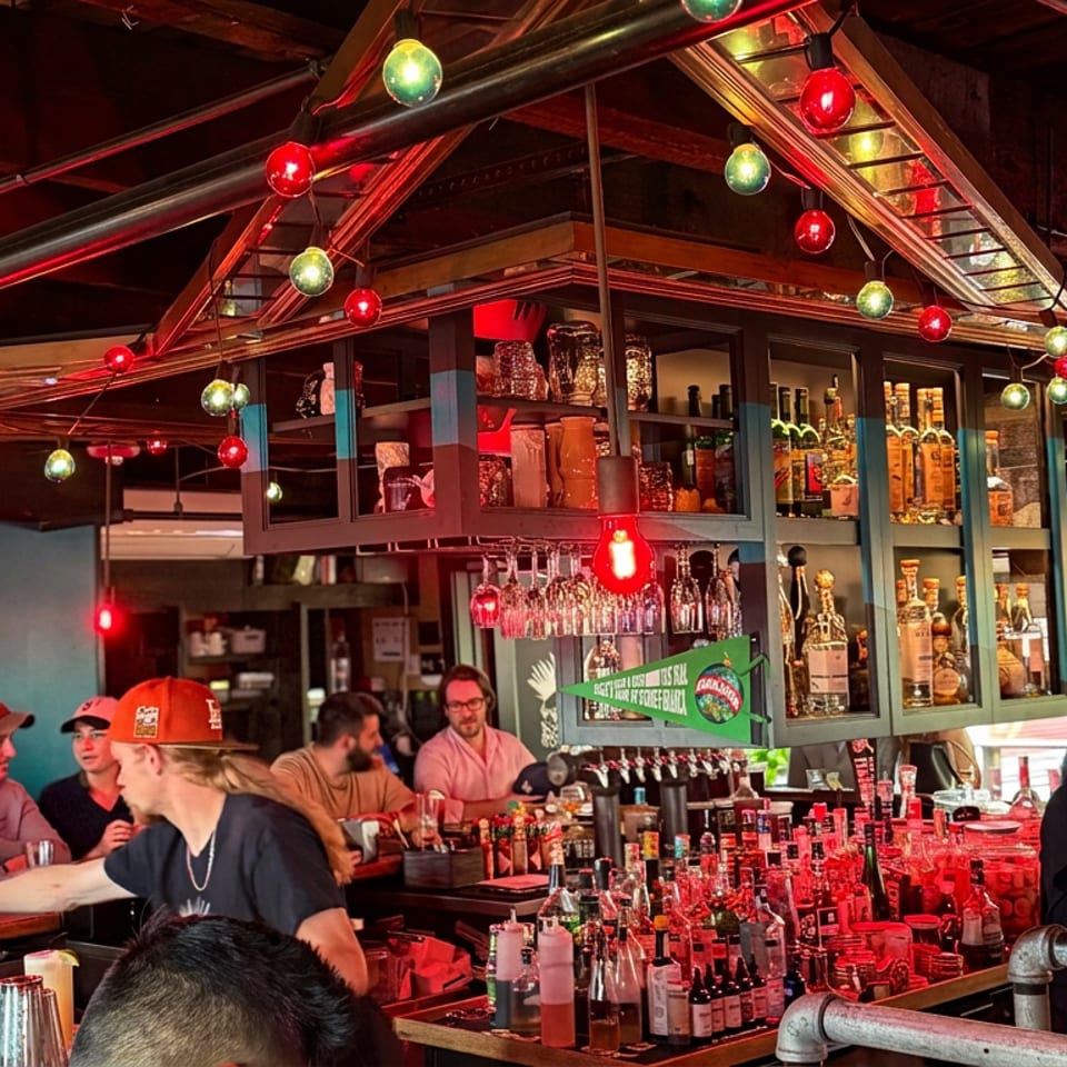 Lively bar interior with colorful string lights, shelves of liquor, bartenders, and patrons gathered around the counter.