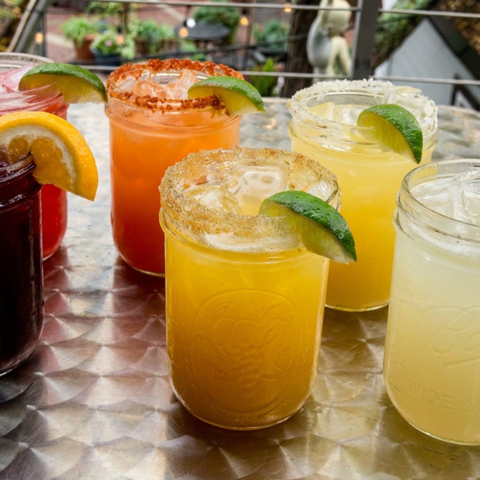 Six colorful cocktails with citrus, cucumber, and chili-salt garnishes arranged on a reflective outdoor restaurant table.