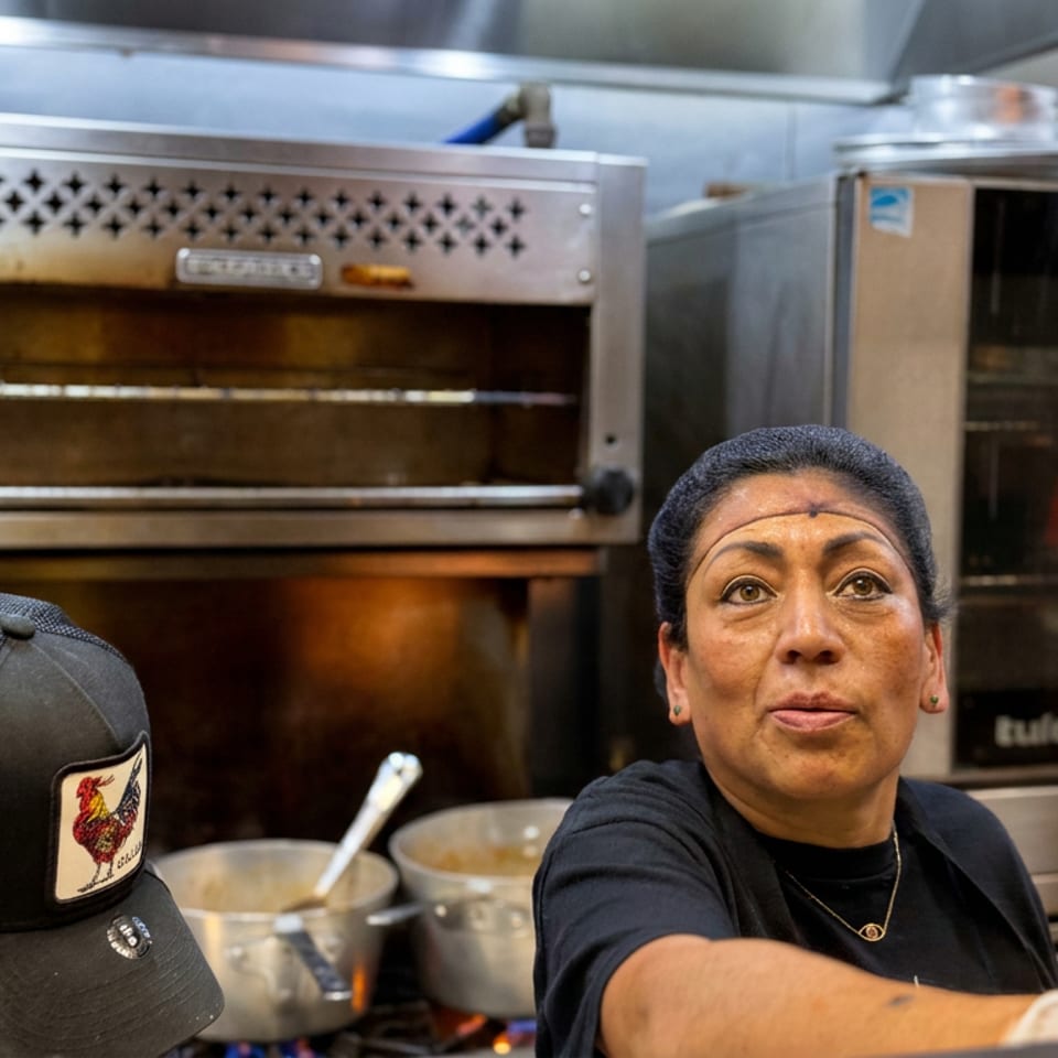 Restaurant kitchen workers prepare food beside commercial ovens, pots, and stainless-steel equipment.