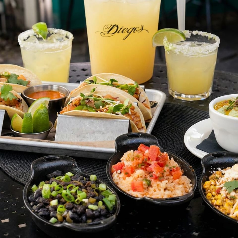 Mexican meal with tacos, black beans, rice, esquites, queso dip, lime wedges, and Diego’s margaritas on an outdoor table.